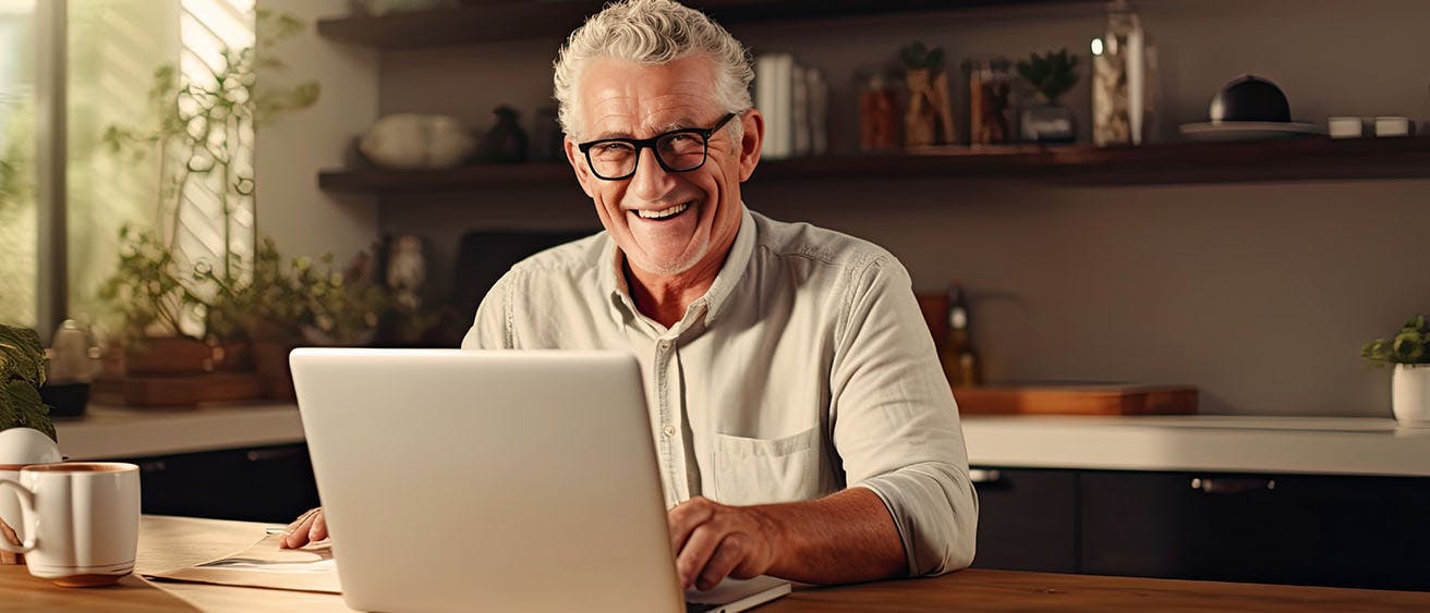 Older man sitting in his kitchen in front of laptop laughing.